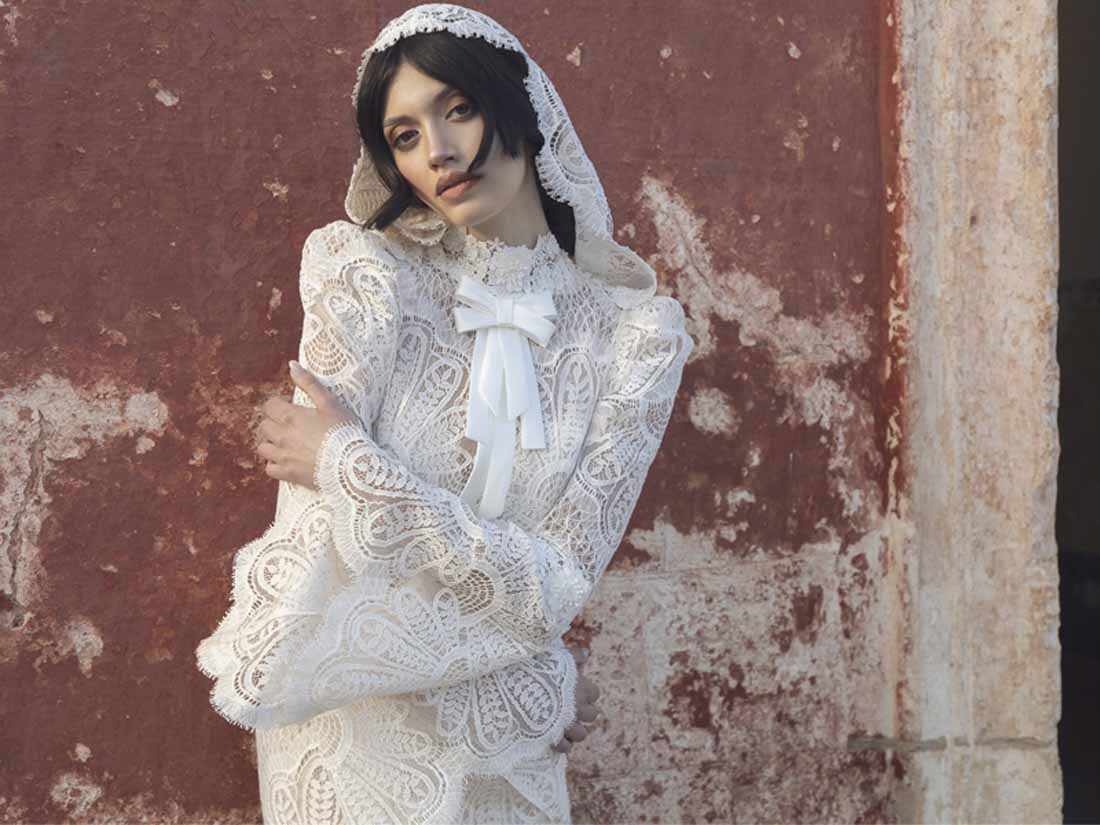 Abito da sposa Pietro De Mita in pizzo con cappuccio e fiocco, contro un muro rosso